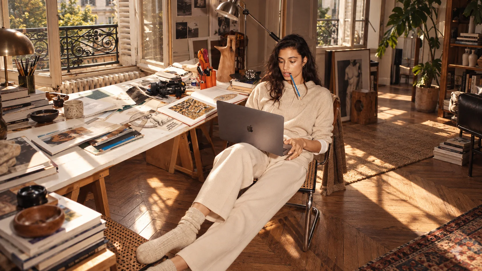 A woman leaning back in a chair with a laptop and a pen in her mouth in a sunlit studio full of prints, books and plants