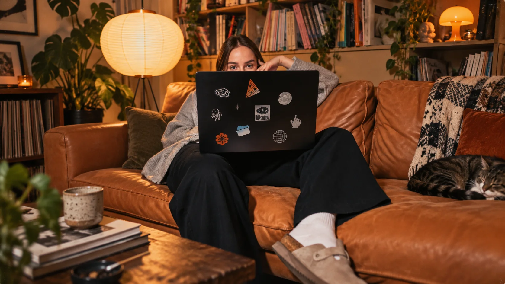 A woman curled up on a leather sofa peeking over a sticker covered laptop, a cat asleep beside her and a paper lamp glowing behind