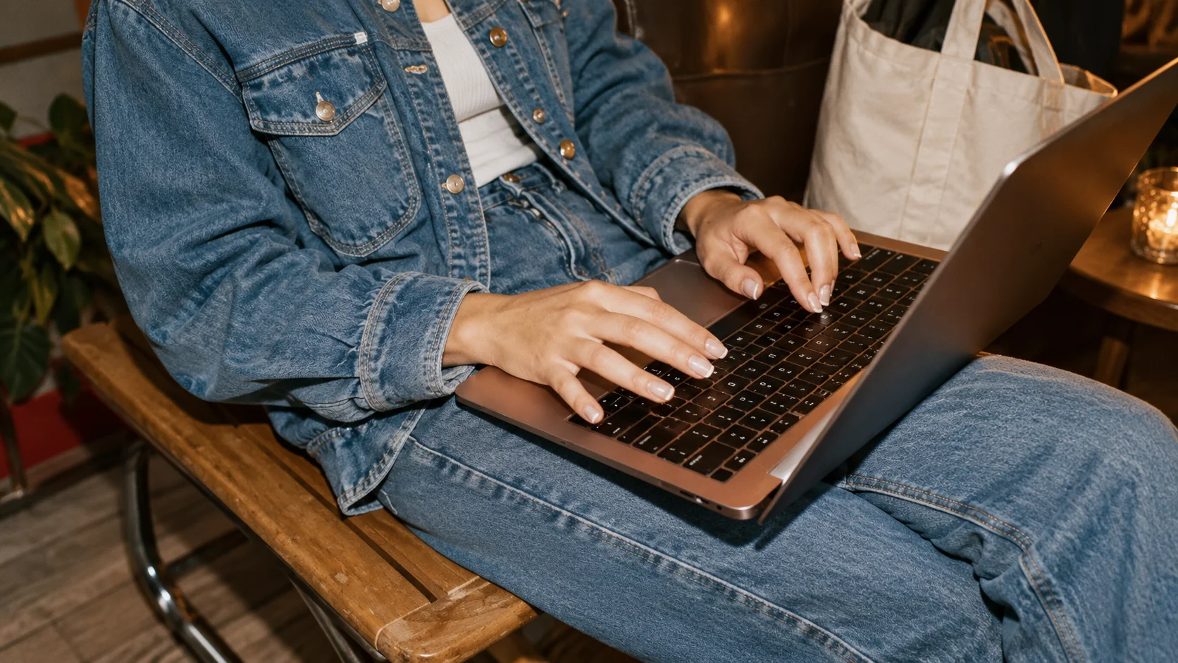 Close up of hands typing on a laptop resting on denim covered knees, a tote bag and candle in the background