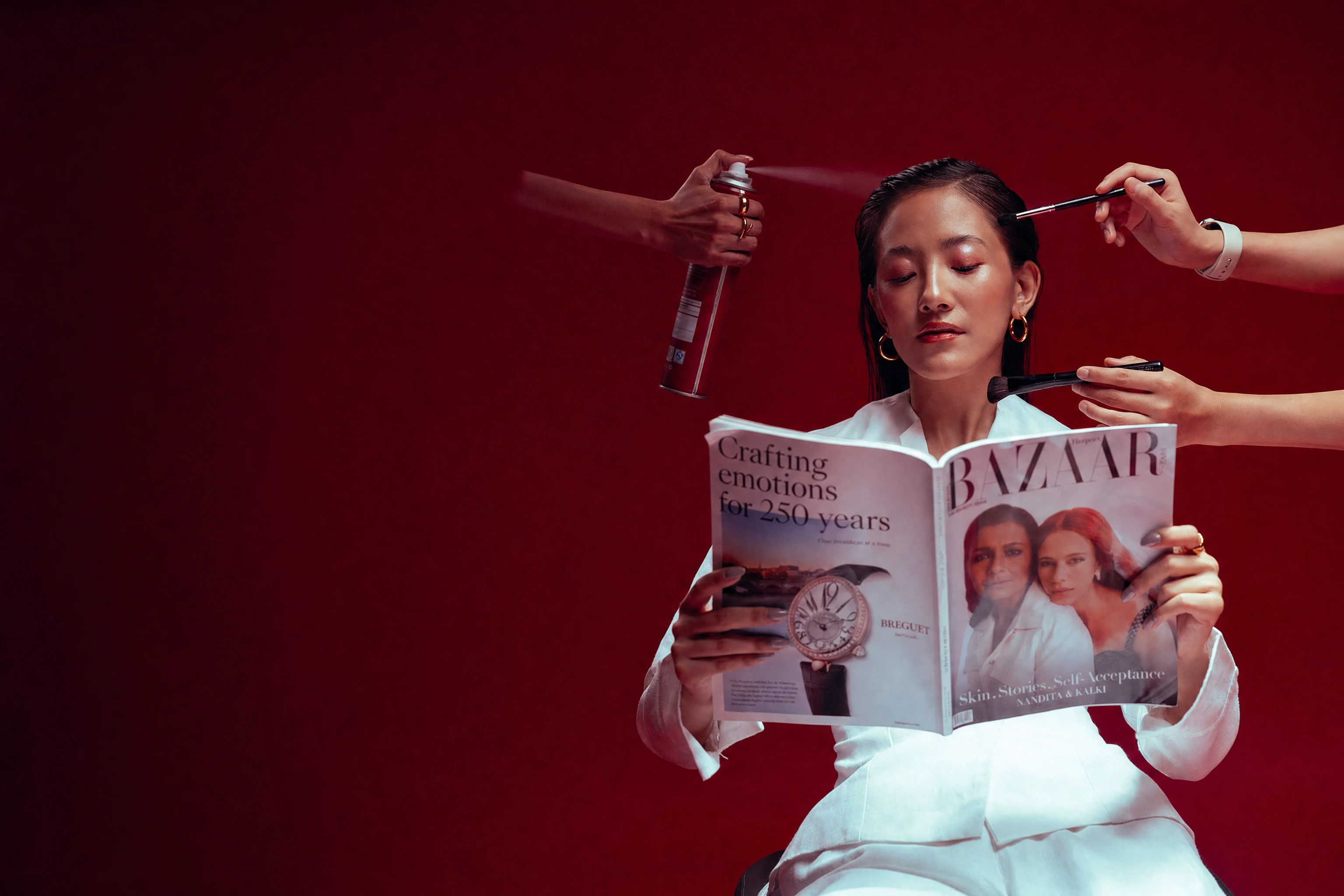 A woman reading a magazine while stylists finish her hair and makeup, on a deep red set