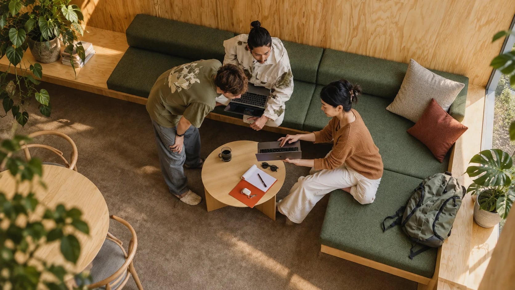 High angle view of three people sharing laptops on a green lounge sofa around a small round table in a plywood room