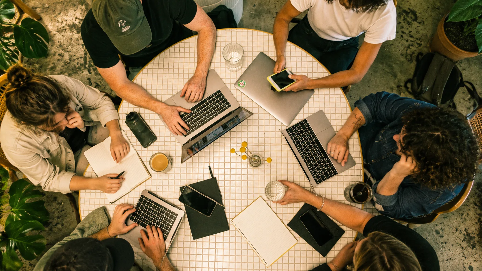 Overhead view of six people working together around a round tiled table with laptops, notebooks, coffee and a phone